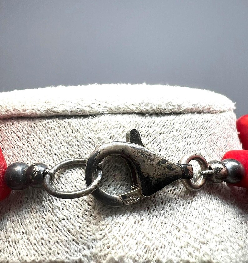May include: Close-up of a silver necklace clasp with a lobster claw closure. The clasp is connected to a strand of red beads. The clasp is stamped with "925". The necklace is resting on a textured, light gray surface.