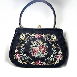 Könnte beinhalten: Schwarze Handtasche mit floralem Kreuzstich-Design. Die Tasche hat einen schwarzen Griff und einen goldenen Verschluss. Das Kreuzstich-Design umfasst rosa, weiße und gelbe Blumen mit grünen Blättern.