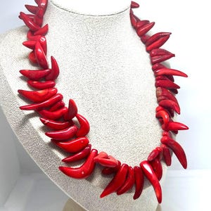 May include: A vibrant red coral necklace displayed on a mannequin. The necklace features numerous curved, horn-shaped pieces of coral, strung together to form a statement piece. The bright red color of the coral stands out against the neutral background.