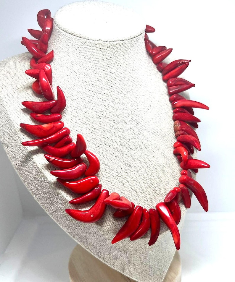 May include: A vibrant red coral necklace displayed on a mannequin. The necklace features numerous curved, horn-shaped pieces of coral, strung together to form a statement piece. The bright red color of the coral stands out against the neutral background.