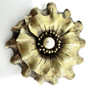 Puede incluir: Una flor decorativa de metal de estilo antiguo, en tonos dorados y bronce. La flor tiene un diseño detallado de pétalos y una perla central rodeada de pequeñas cuentas oscuras. Una pieza ornamental única.