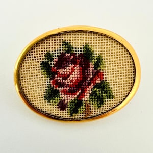 Könnte beinhalten: Eine ovale Brosche im Vintage-Stil mit einem goldfarbenen Rahmen und einem handgestickten Blumendesign. Das Design zeigt eine rosa und violette Rose mit grünen Blättern auf einem beigefarbenen Hintergrund.