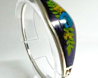 Puede incluir: Pulsera articulada plateada con incrustación de esmalte morado oscuro. La incrustación presenta un diseño floral con detalles en azul, amarillo y verde. La pulsera se exhibe sobre un soporte transparente, mostrando su forma curva y detalles intrincados.