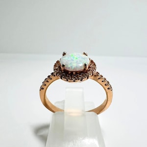 Könnte beinhalten: Roségoldener Ring mit einem großen, ovalen Opal-Edelstein, umgeben von einem Halo aus kleineren Steinen. Das Band ist ebenfalls mit kleinen Akzentsteinen verziert, was einen funkelnden Effekt erzeugt. Der Ring wird auf einem weißen Ständer präsentiert.