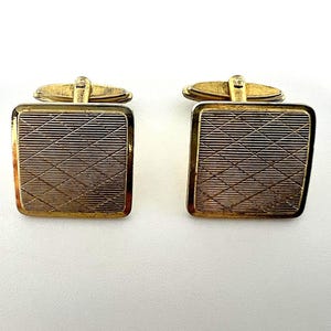 May include: A pair of gold-toned cufflinks. Each cufflink has a rectangular face with a textured, cross-hatched pattern. The edges are framed in gold, and the backing has a hinged clasp for securing to a shirt cuff. The cufflinks are displayed on a white background.