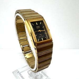 Könnte beinhalten: Eine goldfarbene Puma Quartz-Uhr mit einem rechteckigen schwarzen Zifferblatt. Die Uhr hat ein goldenes Armband und das Puma-Logo. Auf dem Zifferblatt stehen die Worte "Puma Quartz" und "Water Resist".