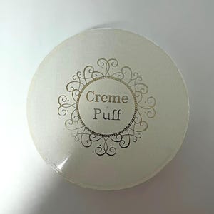 Könnte beinhalten: Ein rundes, cremeweißes Kosmetik-Puderetui mit perlmuttartigem Glanz. Der Deckel ist mit einem goldenen, verzierten, kreisförmigen Filigranrahmen versehen, der den Text "Creme Puff" in einer einfachen, dunklen, serifenlosen Schrift umschließt.