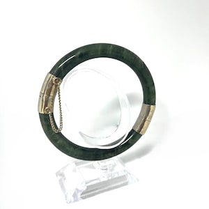 Könnte beinhalten: Ein dunkelgrünes Jade-Armreif-Armband mit silberfarbenen Metallakzenten. Das Armband hat einen Klappverschluss mit einer Sicherheitskette. Der Jade hat eine polierte, glänzende Oberfläche. Das Armband wird auf einem transparenten Ständer präsentiert.