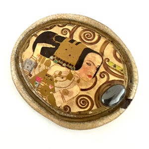 Puede incluir: Broche ovalado con un retrato estilizado de una mujer con cabello negro, labios rojos y un tocado decorativo. La obra de arte está sobre un fondo beige arremolinado con un acento de piedra gris oscuro.