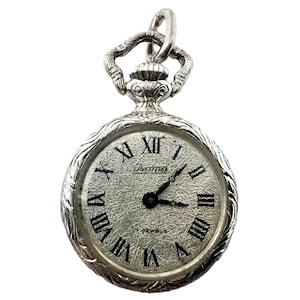 May include: A silver pocket watch with a textured face and black Roman numeral hour markers. The watch face displays the brand name "Isoma" and "17 Jewels". The watch has a decorative loop for hanging.