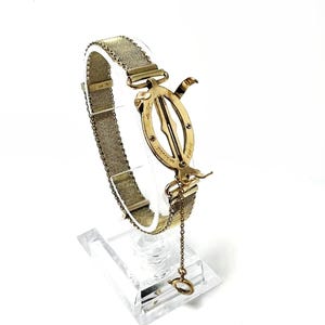 May include: A vintage gold-toned mesh bracelet with an ornate clasp. The clasp features an oval design with intricate details and a small chain with a ring at the end. The bracelet is displayed on a clear stand against a white background.