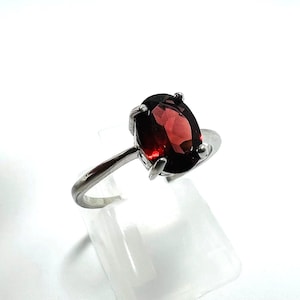 Op de afbeelding: Een zilveren ring met een grote, ovaalvormige, dieprode edelsteen. De edelsteen wordt vastgehouden door vier klauwen. De ringband heeft een eenvoudig, gepolijst zilveren ontwerp, wat een klassieke en elegante uitstraling geeft.