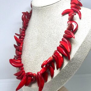 May include: A vibrant red coral necklace displayed on a mannequin. The necklace features a series of polished, claw-shaped coral pieces strung together. The coral has a glossy finish, and the necklace is presented against a neutral background.