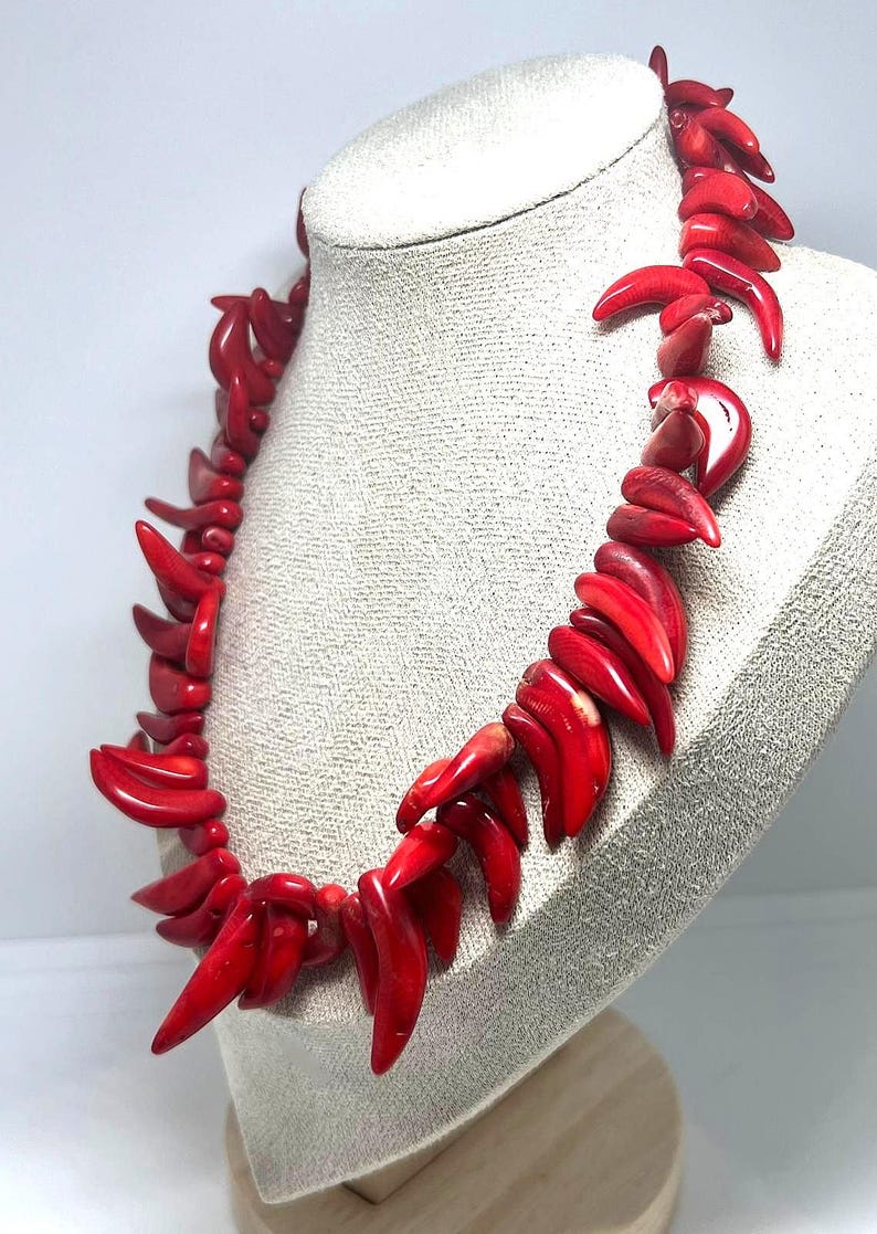 May include: A vibrant red coral necklace displayed on a mannequin. The necklace features a series of polished, claw-shaped coral pieces strung together. The coral has a glossy finish, and the necklace is presented against a neutral background.