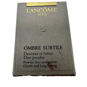 Könnte beinhalten: Eine graue Lidschatten-Duo-Box mit goldenen Buchstaben, die "LANCOME PARIS" lauten. Die Box enthält auch die Wörter "OMBRE SUBTILE" und "Powder duo eyeshadow Gentle and long-lasting".