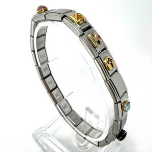 Puede incluir: Una pulsera de eslabones de acero inoxidable con detalles dorados y piedras preciosas. La pulsera presenta eslabones rectangulares, algunos con dijes dorados y piedras preciosas de colores. La pulsera se exhibe sobre un soporte transparente.