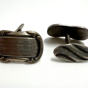 May include: A pair of antique silver cufflinks. One cufflink is rectangular with horizontal lines, framed by a decorative border. The other cufflink is oval with a wavy design. The cufflinks are on a white background.