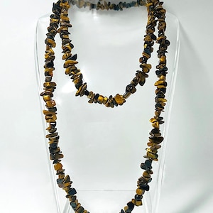 May include: A long necklace and a matching bracelet made of small, irregularly shaped tiger's eye stones. The stones are a mix of brown, gold, and dark blue hues, displayed on a clear stand against a white background.