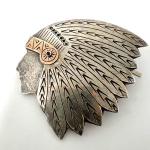 Könnte beinhalten: Eine silberfarbene Metallbrosche in Form eines Personenprofils mit Federschmuck. Der Kopfschmuck weist gravierte Federdetails und ein kupferfarbenes Band mit einem kleinen, runden, dunkelroten Stein in der Mitte auf.