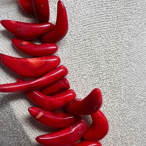 May include: A close-up of a necklace featuring vibrant red coral beads. The beads are smooth and polished, with a curved, organic shape. The necklace is displayed against a neutral, textured background, highlighting the rich color and texture of the coral.