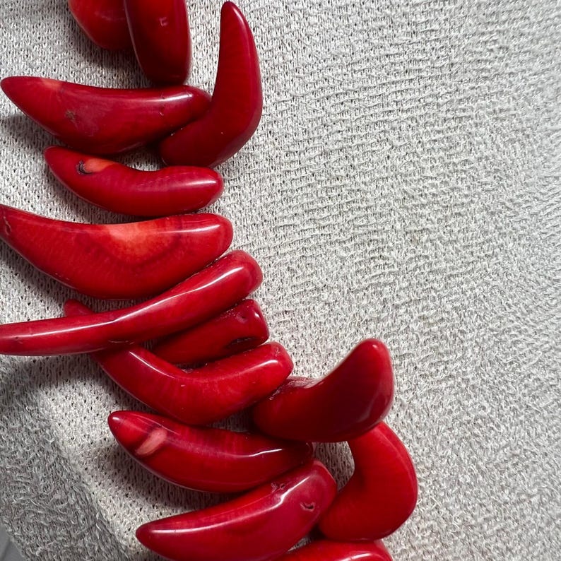 May include: A close-up of a necklace featuring vibrant red coral beads. The beads are smooth and polished, with a curved, organic shape. The necklace is displayed against a neutral, textured background, highlighting the rich color and texture of the coral.