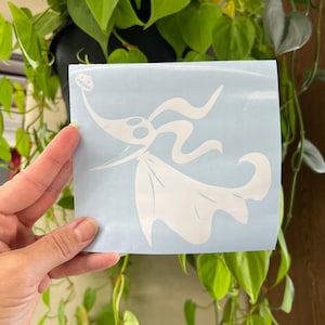 Op de afbeelding: Een witte vinyl sticker met het personage Zero uit The Nightmare Before Christmas. De sticker staat op een lichtblauwe vierkant en wordt voor een achtergrond van groene bladeren gehouden.