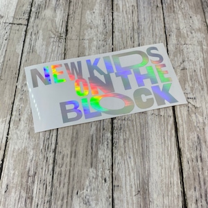 May include: A silver holographic sticker with the text "NEW KIDS ON THE BLOCK" in a rainbow color scheme.
