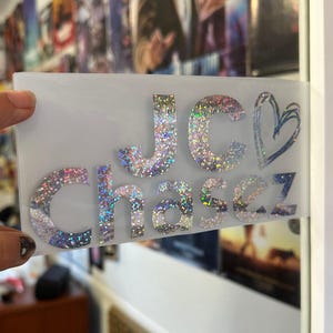May include: A clear sticker with the words "JC Chasez" in glittery, iridescent letters and a blue heart outline. The sticker is held by a hand, with a dark nail polish. The background is blurred.