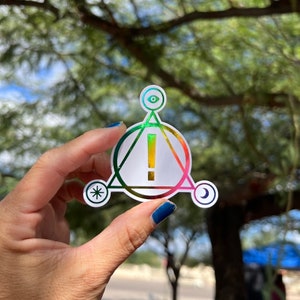 Könnte beinhalten: Ein holografischer Aufkleber mit einem Regenbogen-Farbschema. Der Aufkleber zeigt ein weißes Dreieck mit einem Ausrufezeichen darin, umgeben von einem Kreis. Der Kreis ist von einem größeren Dreieck mit einer Mondsichel, einem Auge und einem Stern an jeder Spitze umgeben.
