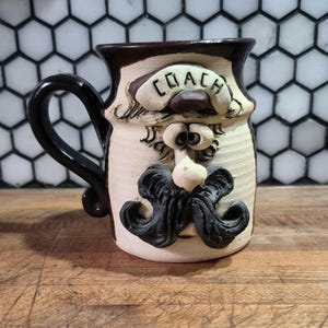 May include: A ceramic mug with a sculpted face design. The mug features a black handle and a brown rim. The face has a mustache, nose, and the word "COACH" on a banner. The mug is cream-colored with black and brown accents.