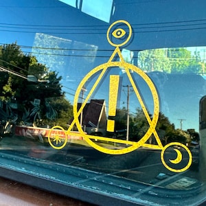 May include: A yellow decal of a stylized triangle with an exclamation point inside a circle. The triangle has an eye at the top, a star at the bottom left, and a crescent moon at the bottom right.