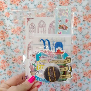 May include: A clear plastic bag filled with colorful stickers and a button. The stickers feature various designs, including a cactus, a bottle of tequila, a sombrero, and a woman with a heart. The button is black with white text that reads "May the Fourth Be With You".