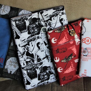 May include: A collection of fabric book covers featuring Star Wars characters and imagery. The covers display various designs, including Darth Vader, Stormtroopers, and BB-8, in black, white, red, and blue. Some covers have button closures.