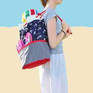 May include: A blue and white striped drawstring bag with a red trim and anchor print. The bag is filled with flip flops and a colorful beach towel. The bag is being worn as a backpack by a person wearing a grey shirt and white skirt.