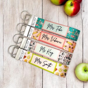 May include: Four keyrings with silver hardware and patterned fabric straps. Each strap displays a different design and the names "Mrs. Feke", "Mrs. Vukovic", "Ms. King", and "Mrs. Smith". Apples are visible on a wooden surface.