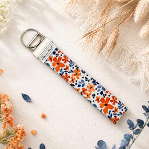 May include: A floral wristlet key fob with a silver-tone metal clasp. The fabric features a white background with orange and blue flower patterns. The text "Robin's Nest of Kensington" is printed on the metal clasp.