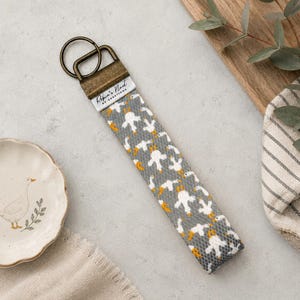 May include: A fabric keyring featuring a repeating pattern of white geese with orange beaks and feet on a grey background. The strap is attached to a bronze-toned metal hardware clasp with a ring, labelled with the text "Ripin's Nest of Creations".