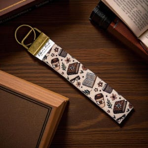 May include: A beige fabric keychain wristlet with a gold-coloured metal clasp. The wristlet features a repeating pattern of books, coffee cups, glasses, and floral elements. The text "Robin's Nest of Creations" is printed on a white label.