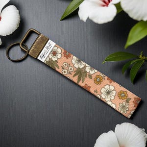 May include: A floral keychain wristlet with a peach background and white, orange, and brown flowers. The wristlet has a bronze-coloured metal clasp and key ring. The text "Robin's Nest of Creations" is printed on a white label.