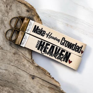 May include: Two beige keychains with the text "Make Heaven Crowded" in black script. The keychains have bronze-colored hardware and a small red heart. The brand name "Robin's Nest of Creations" is printed on the side.