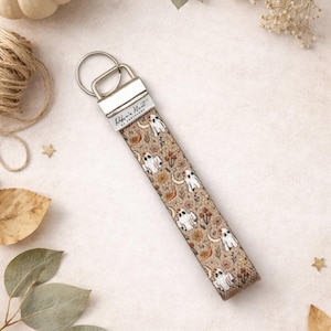 May include: A beige fabric wrist strap keychain with a repeating pattern of white ghosts, crescent moons, and floral elements. The keychain has a silver metal ring and a silver metal clasp with the text "Robin's Nest of Creations".