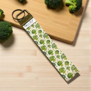 May include: A fabric wristlet key fob with a repeating pattern of green broccoli florets on a cream background. The key fob has a bronze-colored metal clasp and a small white label with the text "Robin's Nest of Creations".