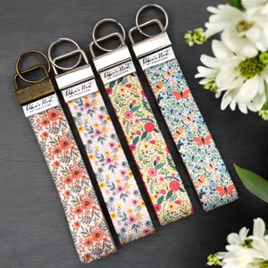 May include: Four key fobs with floral patterns and metal fixings. The key fobs display various floral designs, including orange, purple, and red flowers, and butterflies. Each fob has a small tag that reads "Robin's Nest of Creations".