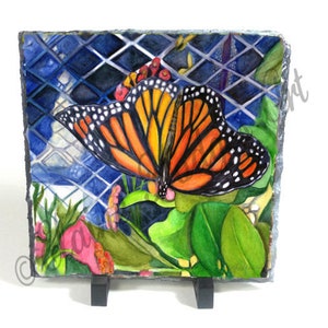 May include: A watercolor painting of a monarch butterfly with orange and black wings, perched on a green leaf with pink flowers. The butterfly is painted on a slate tile with a blue and white grid pattern.