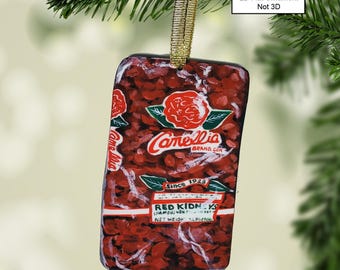 May include: A red and white Christmas ornament shaped like a can of Camelia Brand Red Kidneys. The ornament features a red rose with green leaves and the text "Camelia Brand Con" and "Since 1928".