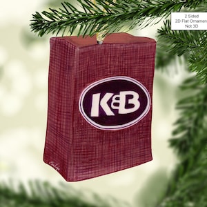 May include: A burgundy-colored Christmas ornament with a white oval containing the text "K&B" in black lettering. The ornament is 2-sided and flat, not 3D.