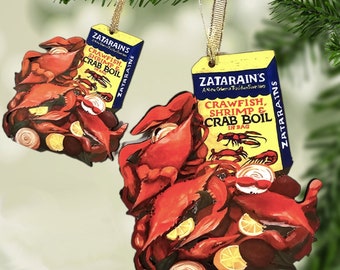 Puede incluir: Dos adornos navideños de madera con forma de cajas de Zatarain's Crawfish, Shrimp & Crab Boil. Las cajas son amarillas con texto azul y rojo. Los adornos presentan una pintura realista de cangrejos de río, camarones, limones y cebollas.
