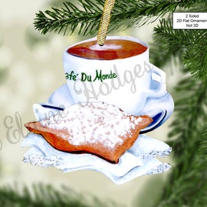 May include: A white ceramic coffee mug with the text "Cafe' Du Monde" in green lettering, filled with brown coffee, and a beignet dusted with powdered sugar on a white napkin. The mug has a gold handle and is hanging from a gold string.