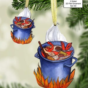 May include: A blue pot with a red crab and other ingredients inside, sitting on a bed of orange flames. The pot is hanging from a gold ribbon and has a white tag that says "2 Sided 2D Flat Ornament Not 3D".