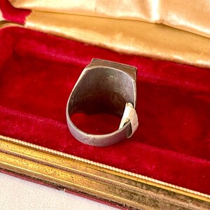 K&ouml;nnte beinhalten: Ein silberfarbener Ring mit rechteckiger Oberseite, pr&auml;sentiert in einer roten Samt-Schmuckschatulle. Der Ring hat ein schlichtes Band und ein leicht abgenutztes Aussehen. Die Schatulle hat einen goldfarbenen Rand und ein cremefarbenes Satinfutter.